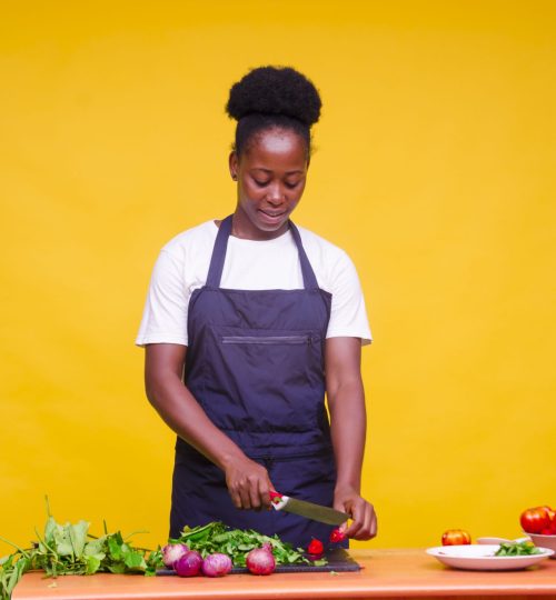 A horizontal shot of a young attractive African cook cutting vegetables with a knife/ orange background